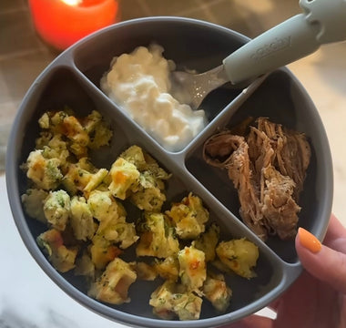 Gray divided baby plate with broccoli bites, yogurt, and shredded meat, held by hand with orange nail polish.