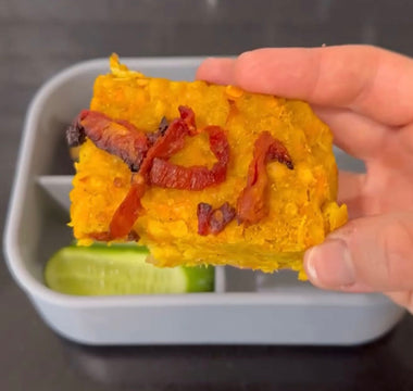 Hand holding savory yellow baked dish with sun-dried tomatoes over bento box with cucumber slices