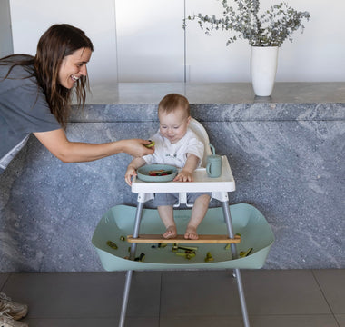 Baby in high chair with mess-free food catcher, mom feeding with smile