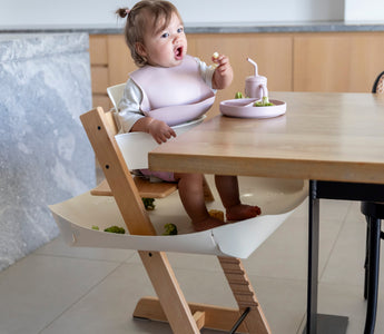 Baby eating with mess-free bib and food catcher under high chair, protecting floors during mealtime.